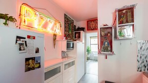 A kitchen with a neon sign reading "PIZZA," a refrigerator with photos, and a microwave.