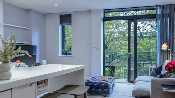 Eine moderne Küche mit weißer Arbeitsplatte, Barhocker und einem Topf mit Pflanze. Ein großes Fenster bietet Blick auf Bäume, und eine Glastür führt in einen Wohnbereich mit Sofa und Hocker.