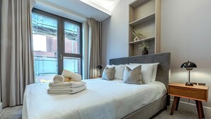 Ein ordentlich bezogenes Bett mit Kissen und einem gefalteten Handtuch, ein Nachttisch mit Lampe und ein Fenster mit Vorhängen.
