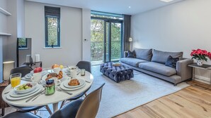 Ein modernes Wohnzimmer mit einem grauen Sofa, einem Esstisch mit Frühstück und einem großen Fenster mit Blick auf Bäume.