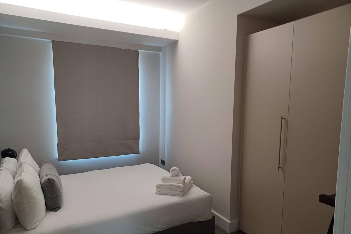 A bedroom with a bed, pillows, a window with blinds, and a wardrobe.