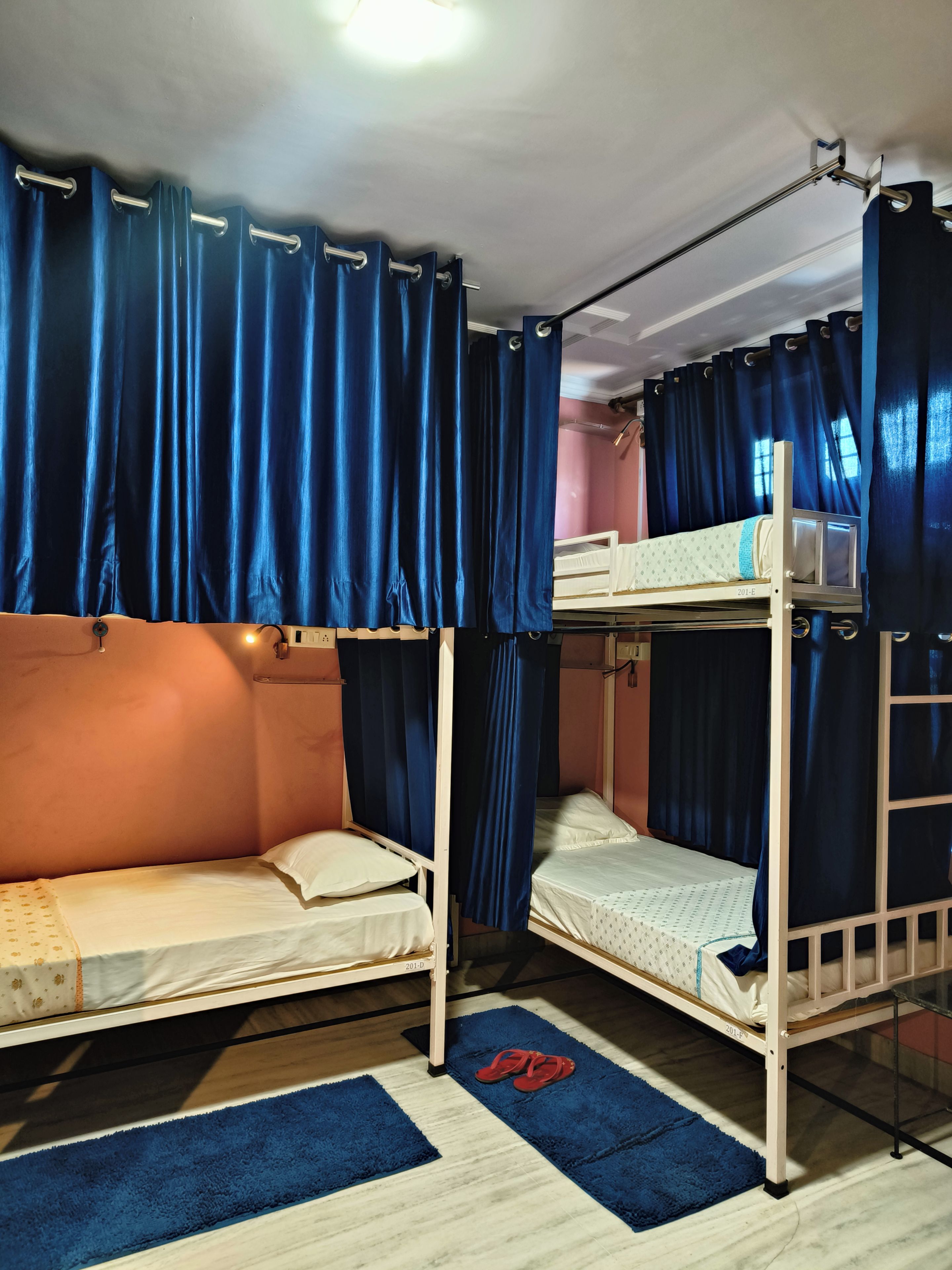 A dormitory room with two bunk beds, blue curtains, a small table, and a pair of red slippers on a blue rug.