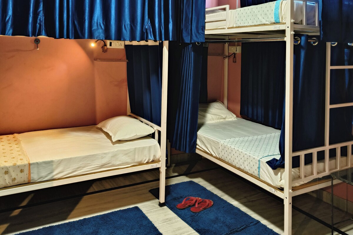 Een studentenkamer met twee stapelbedden, blauwe gordijnen, een klein tafeltje en een paar rode pantoffels op een blauw vloerkleed.