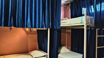 A dormitory room with two bunk beds, blue curtains, a small table, and a pair of red slippers on a blue rug.