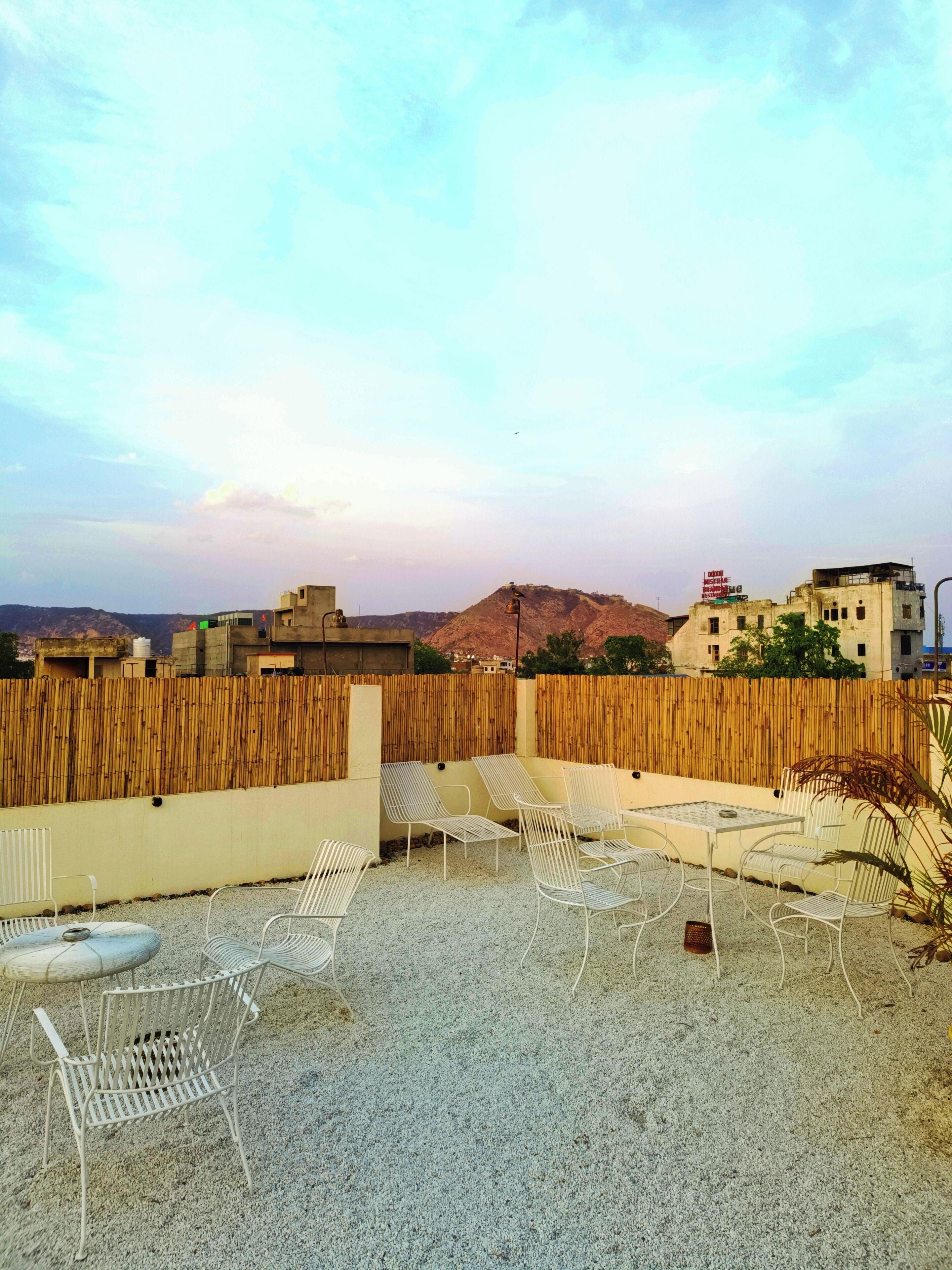 Una terraza en la azotea con sillas y mesas de metal blanco, una valla de bambú y vistas a edificios y colinas a lo lejos.