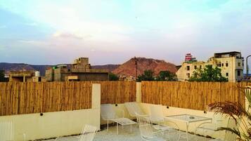 Una terraza en la azotea con sillas y mesas de metal blanco, una valla de bambú y vistas a edificios y colinas a lo lejos.