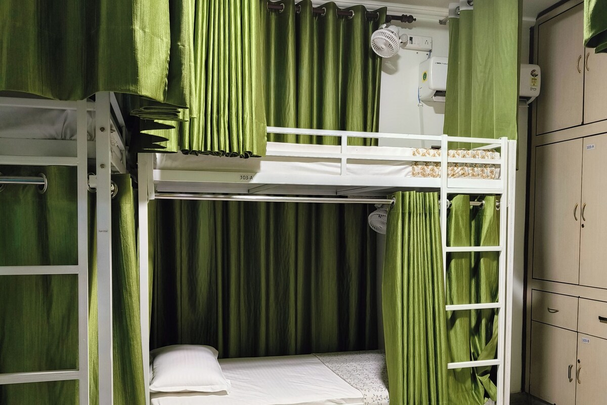 Een slaapkamer met een stapelbed, groene gordijnen, een witte ventilator en een kledingkast.