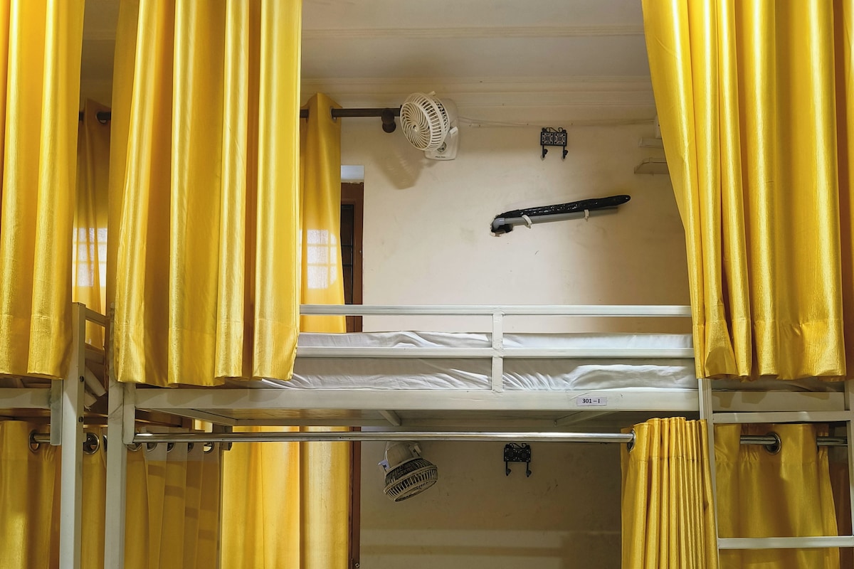 Een slaapkamer met stapelbedden, gele gordijnen, een ventilator en een klein plankje.
