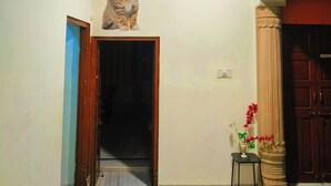 A hallway with a ceiling fan, a door leading to a dark room, a small table with a vase of flowers, and a decorative pillar. - Ohh Hostler Jaipur (Jaipur)