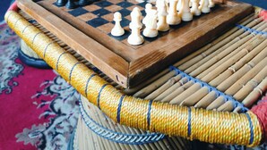 A chessboard on a woven mat with a patterned carpet, a mirror on the wall, and a curtain in the background.