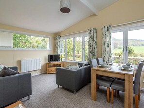 A cozy living room with a sofa, a dining table with chairs, a TV on a stand, and large windows with curtains. - Bridgend Chalet on Reelig Estate (Inverness)