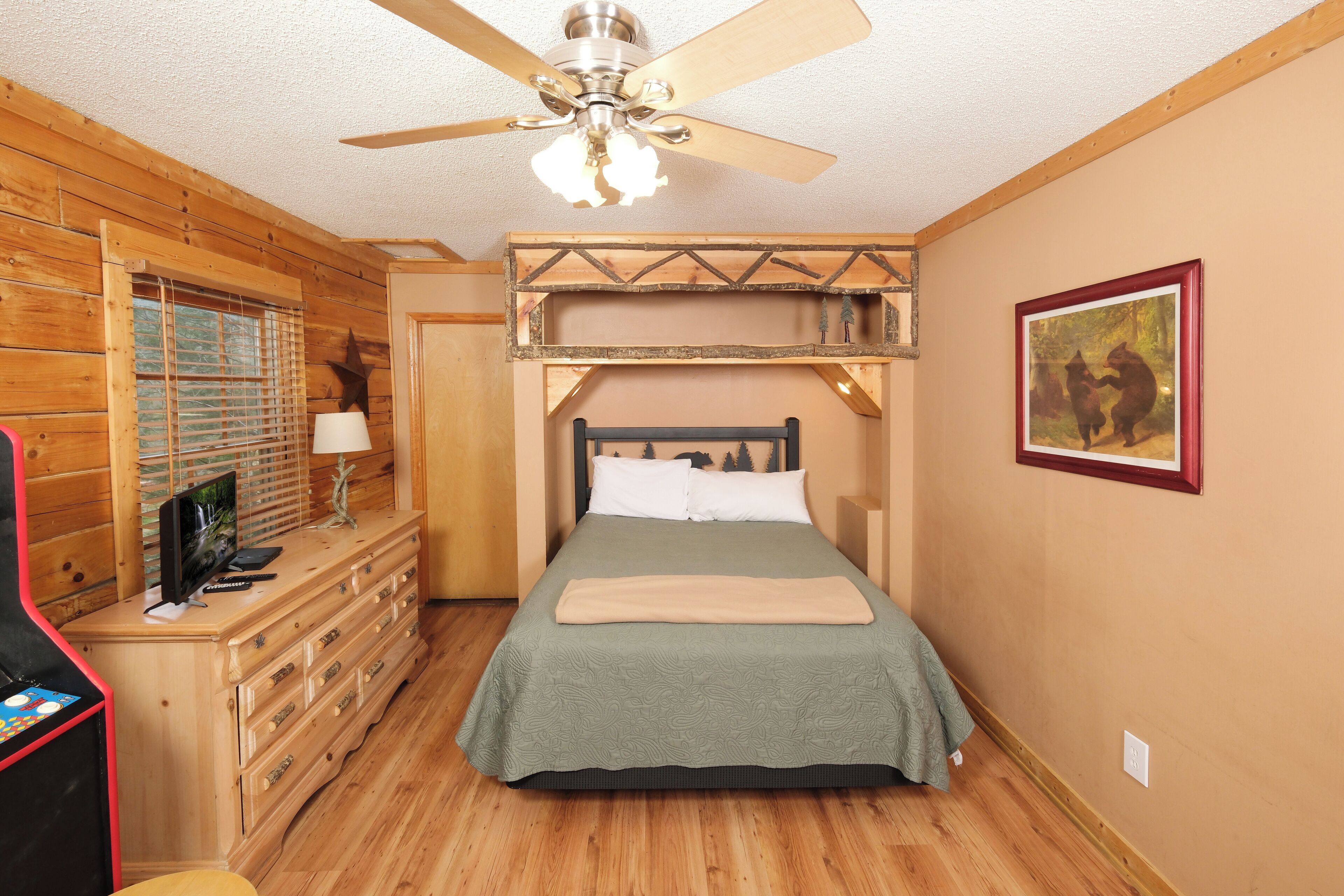 Une chambre à coucher avec un lit, une commode en bois, un ventilateur de plafond et un tableau encadré au mur.