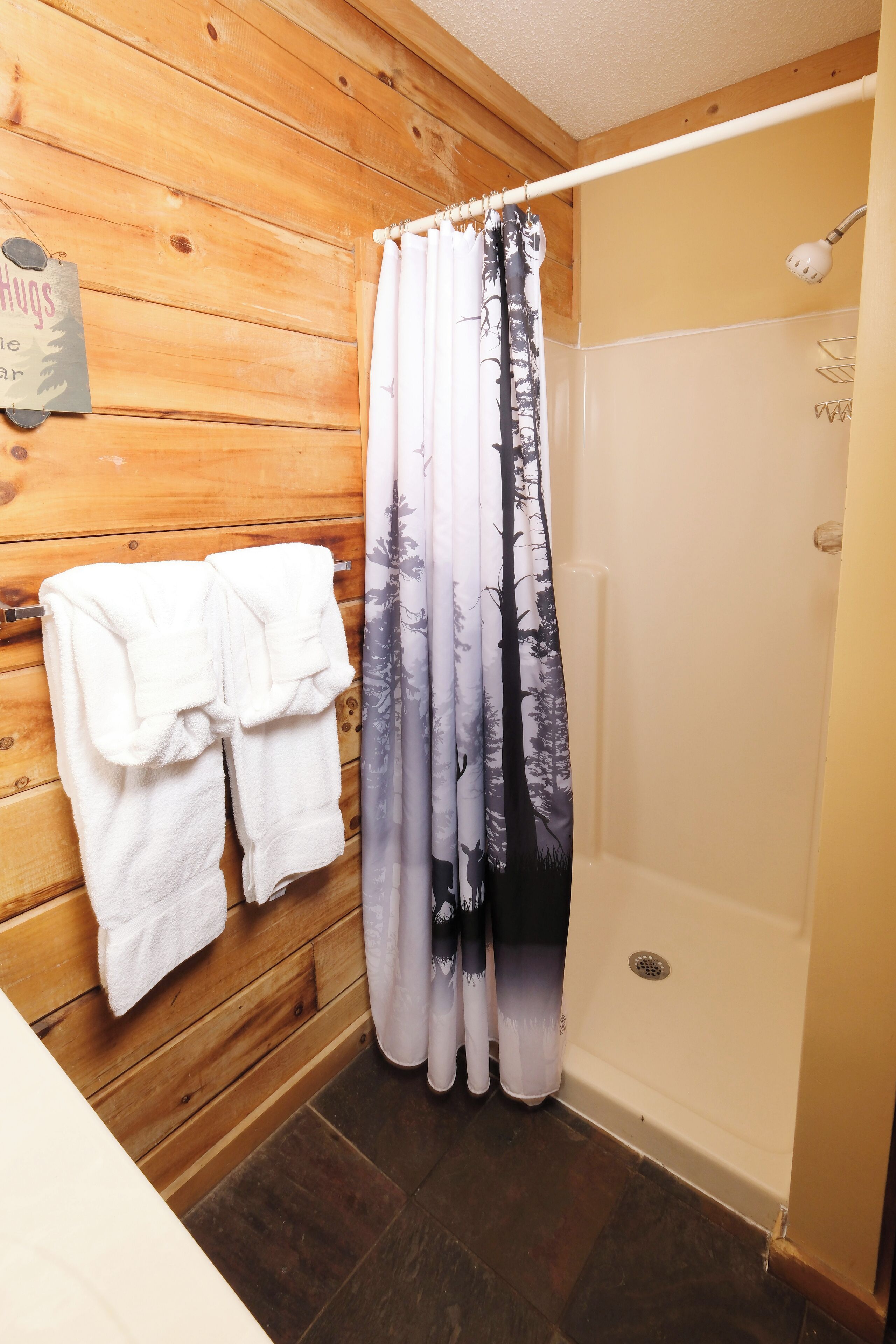 Un baño con paneles de madera y cortina de ducha estampada con un diseño de bosque. Hay dos toallas blancas colgadas en la pared.