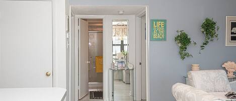 Un couloir avec une porte blanche, une porte en verre donnant sur une salle de bain et un coin salon avec un canapé à motifs.
