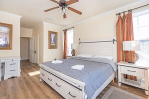 Quarto com cama, mesas de cabeceira, ventilador de teto e janela com cortinas.