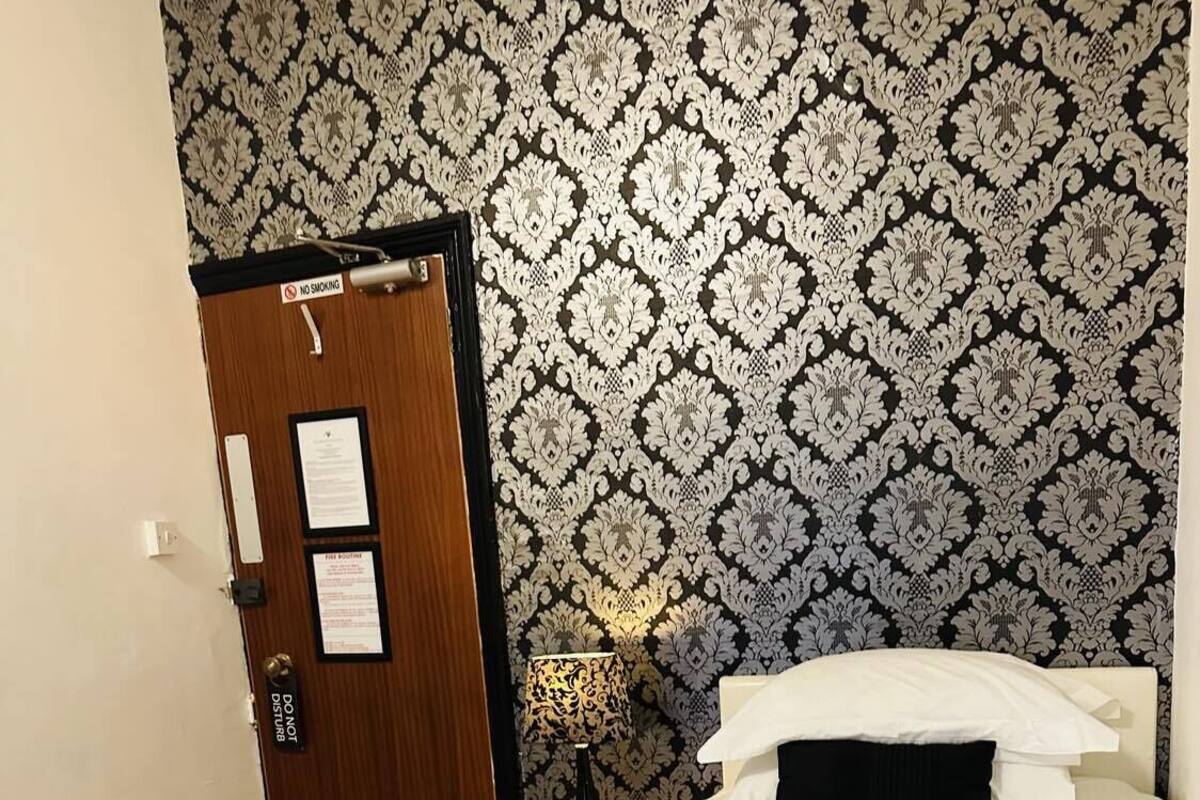 A hotel room with a bed, a nightstand, a lamp, and a door with a 'No Smoking' sign.