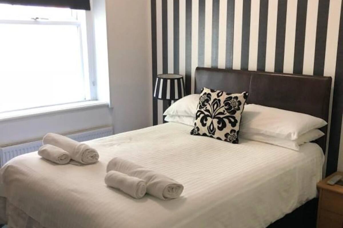 A neatly made bed with white linens and rolled towels, a striped wall pattern, a bedside lamp, and a window with blinds.