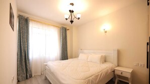 Un dormitorio con una cama grande, una mesita de noche, un aparador, una araña y una ventana con cortinas.