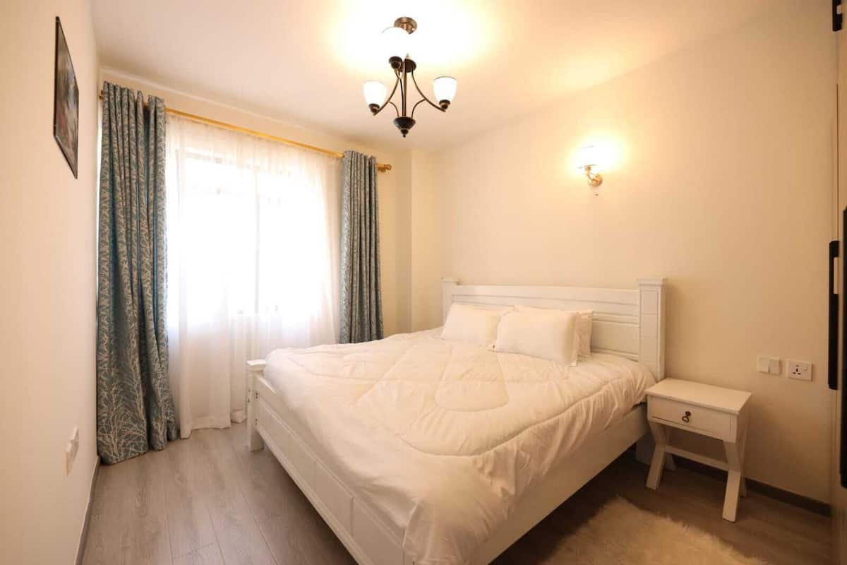 Un dormitorio con una cama grande, una mesita de noche, un aparador, una araña y una ventana con cortinas.