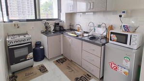 Cocina compacta con estufa a gas, horno, microondas y un refrigerador pequeño. Hay una ventana sobre el fregadero y se ve un cubo de basura.