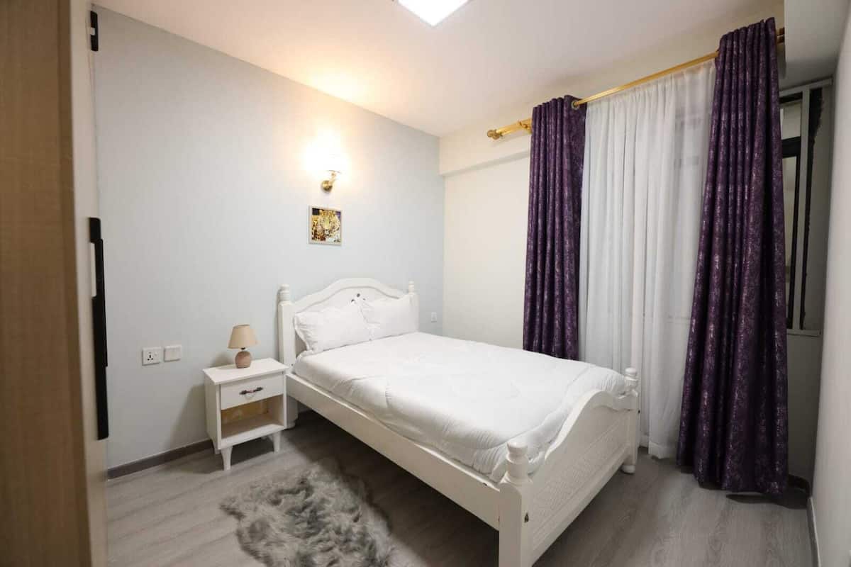 Un dormitorio con una cama blanca, una mesita de noche, una lámpara y una alfombra pequeña.