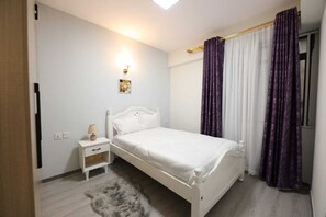 A bedroom with a white bed, a nightstand, a lamp, and a small rug. - Lux Suites Sandalwood  Apartments (Nairobi)