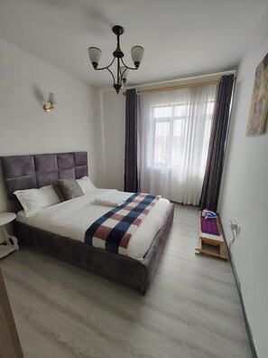 A bedroom with a large bed, a chandelier, a window with curtains, and a small table with a chair. - Lux Suites Sandalwood  Apartments (Nairobi)