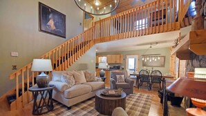 A spacious living area with a wooden staircase, a sofa set, a dining table with chairs, and a kitchen in the background. - Hayden’s Hideaway - View, hot tub, game room, fun! (Blairsville)