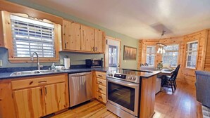 A kitchen with wooden cabinets, a dishwasher, an oven, and a dining area with a table and chairs. - Hayden’s Hideaway - View, hot tub, game room, fun! (Blairsville)