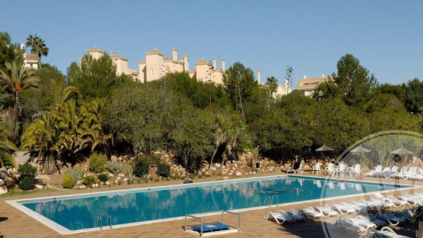 A swimming pool with lounge chairs and umbrellas, surrounded by greenery and a building with multiple chimneys. - La Manga Club Bellaluz Apartment 603 (Atamaría)