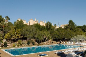 Ein Schwimmbecken mit Liegestühlen und Sonnenschirmen, umgeben von Grünpflanzen und einem Gebäude mit mehreren Schornsteinen.