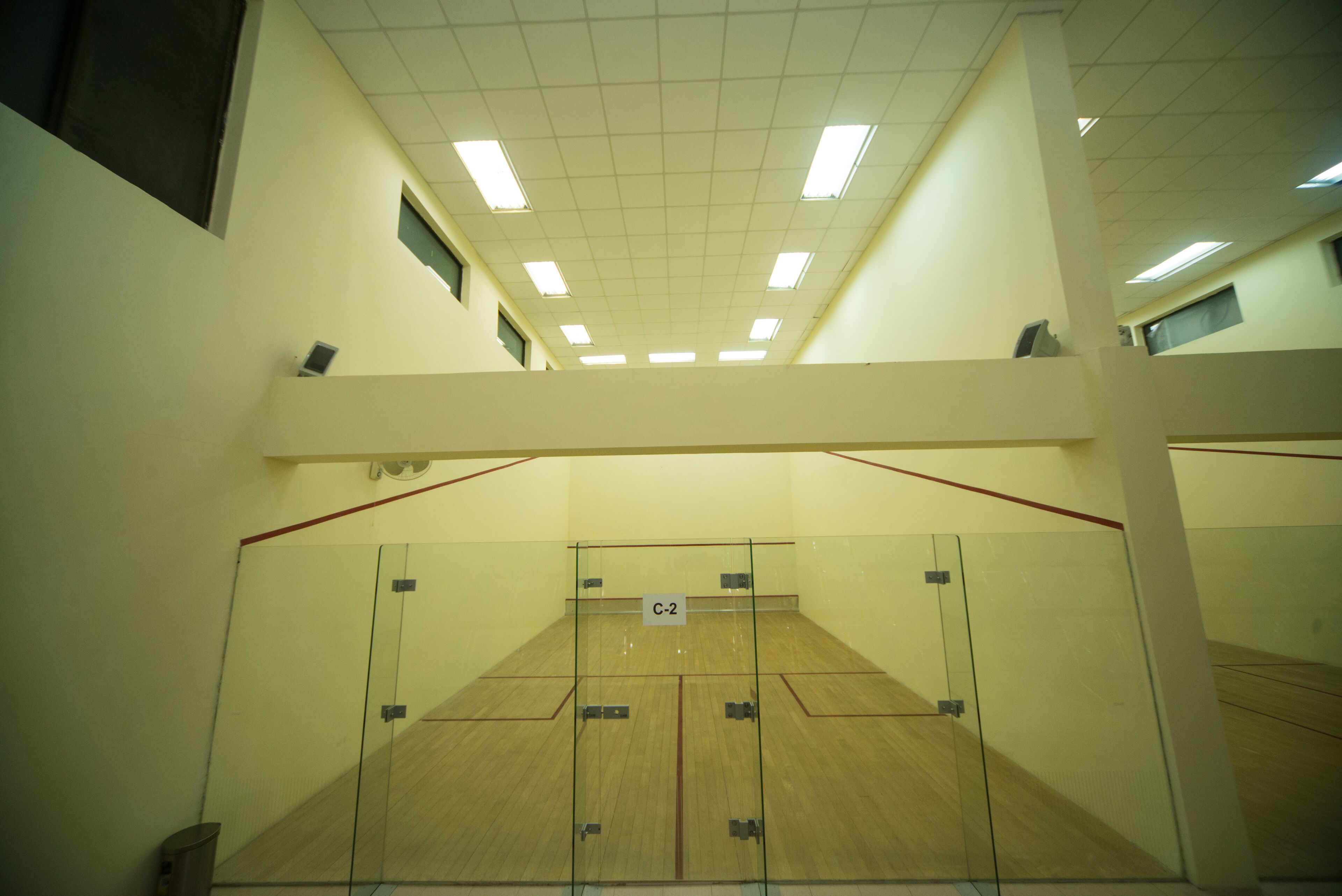 A squash court with glass walls, a wooden floor, and a ceiling with fluorescent lights.