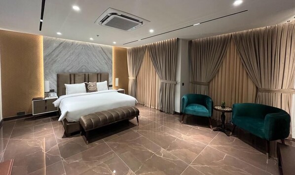 Habitación de hotel moderna con una cama grande, una mesita de noche, un banco, dos sillas y una pequeña mesa.