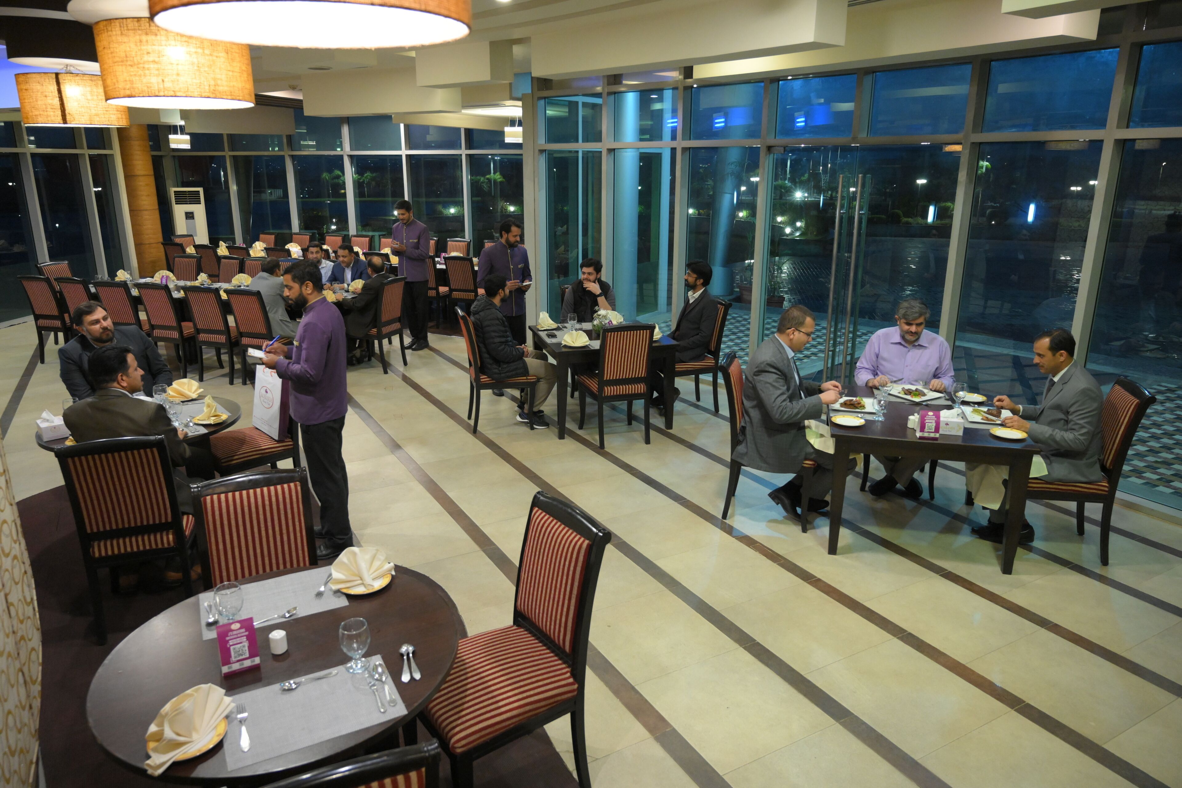 A spacious dining area with multiple tables and chairs, large windows offering a view of the outdoors, and a modern interior design.