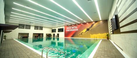 An indoor swimming pool with a 'Dive into Freshness' mural, tiled floors, and overhead lighting.