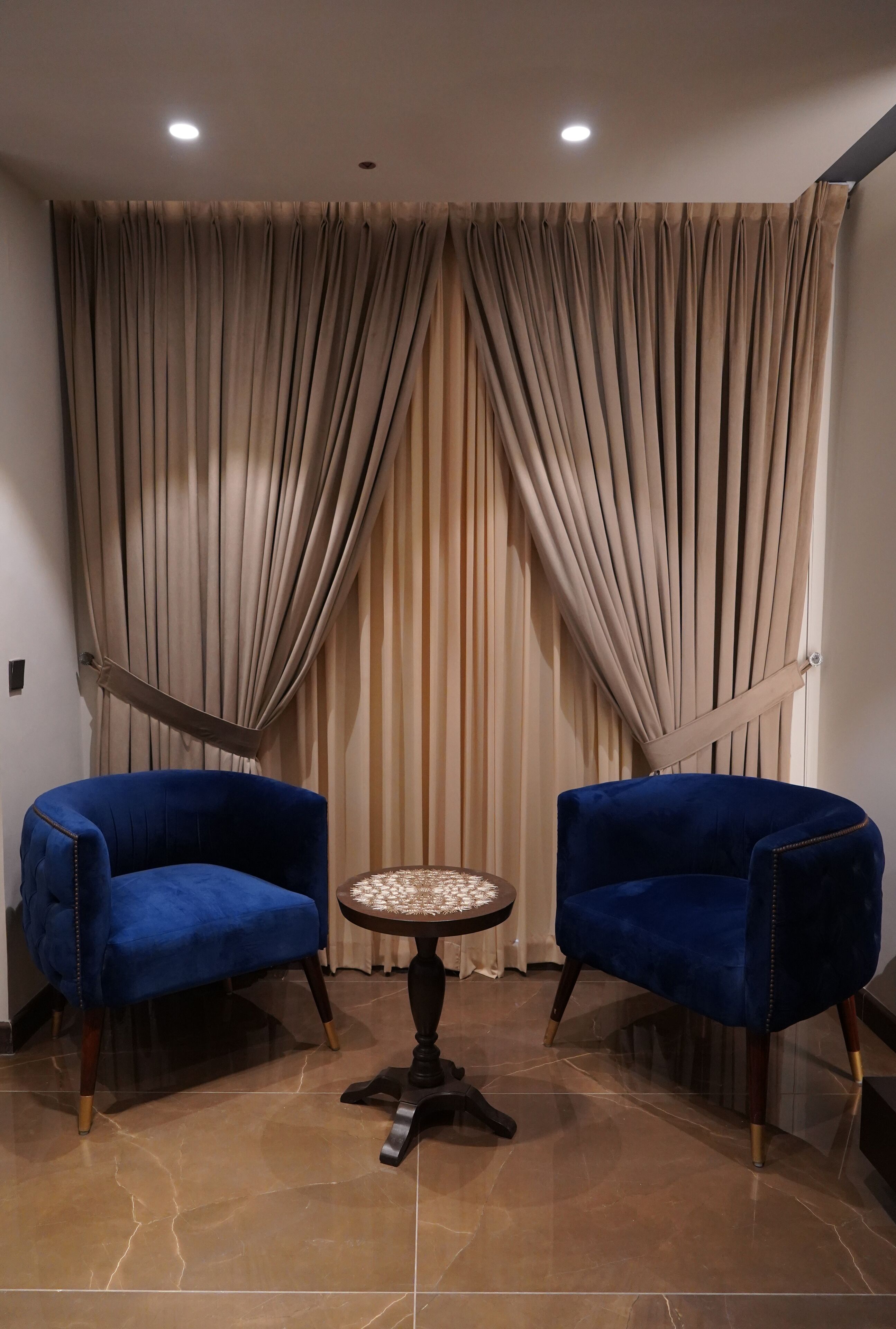 A room with two blue armchairs, a round table, and a large window with beige curtains.