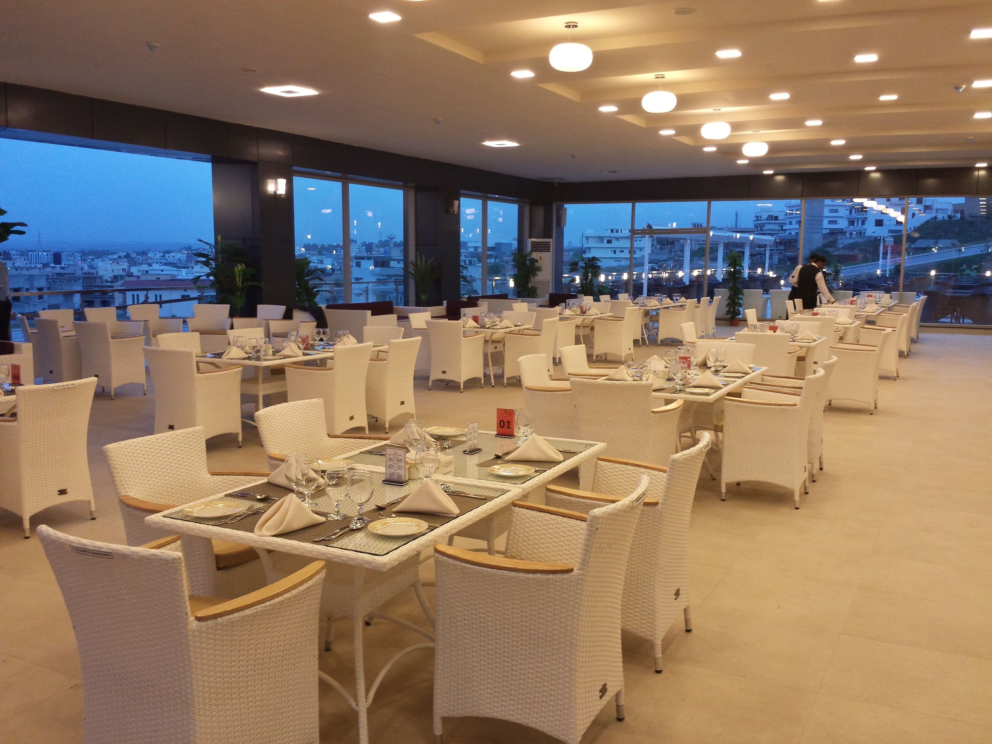 Un restaurant doté de mobilier en osier blanc, de grandes fenêtres offrant une vue sur la ville et d’un intérieur épuré et moderne.