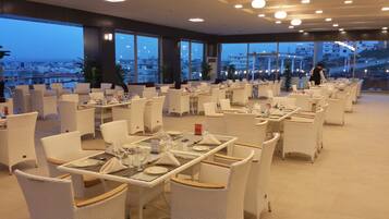 Un restaurant doté de mobilier en osier blanc, de grandes fenêtres offrant une vue sur la ville et d’un intérieur épuré et moderne.