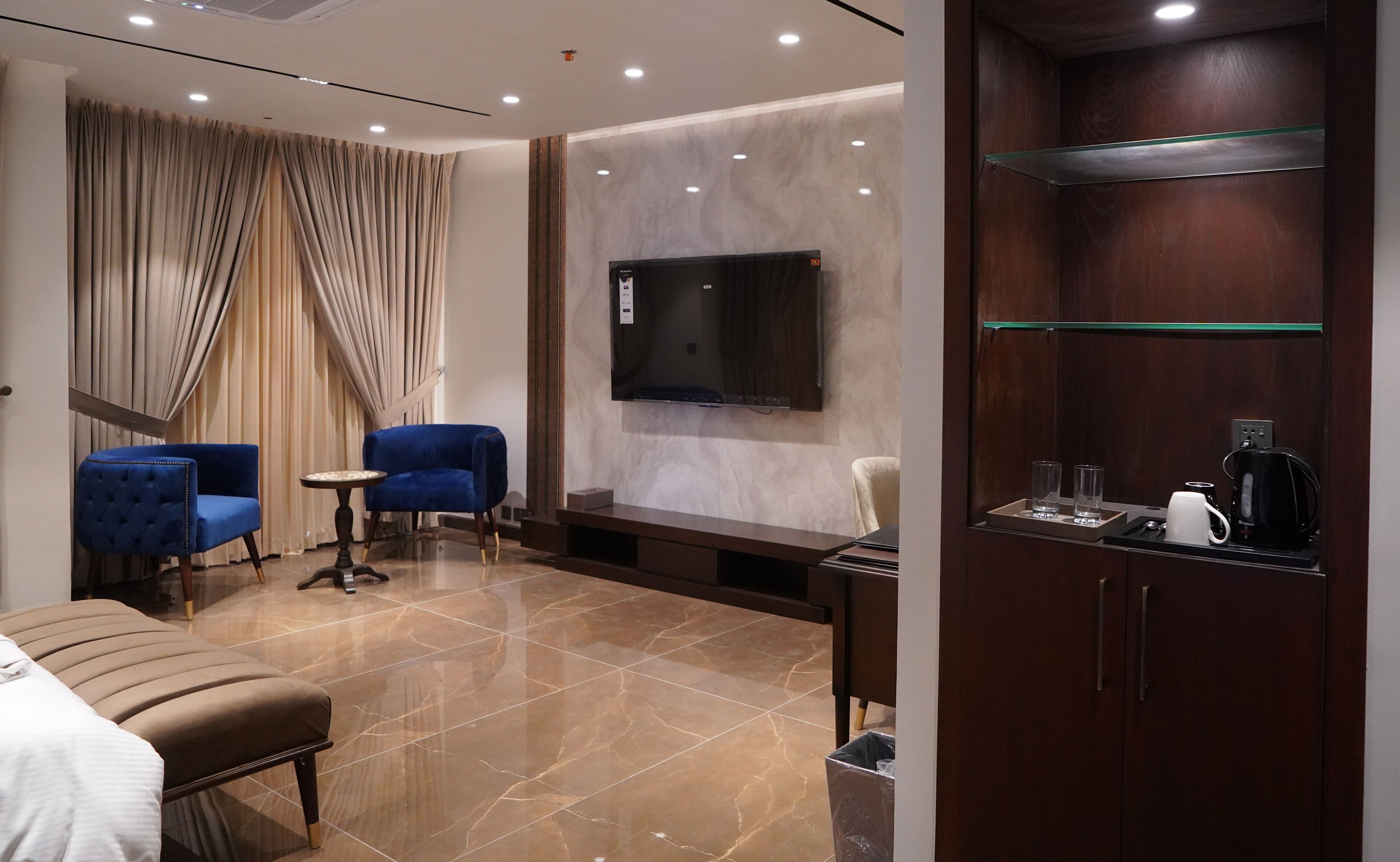 A hotel room with a bed, a blue armchair, a small table, a TV mounted on the wall, a wooden cabinet with a glass shelf, and a coffee maker.