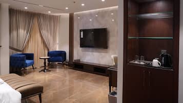 A hotel room with a bed, a blue armchair, a small table, a TV mounted on the wall, a wooden cabinet with a glass shelf, and a coffee maker.