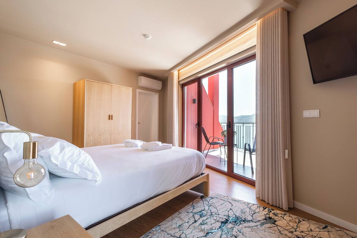 A modern hotel room with a large bed, a flat-screen TV, a balcony with a red wall, and a view of the outdoors.
