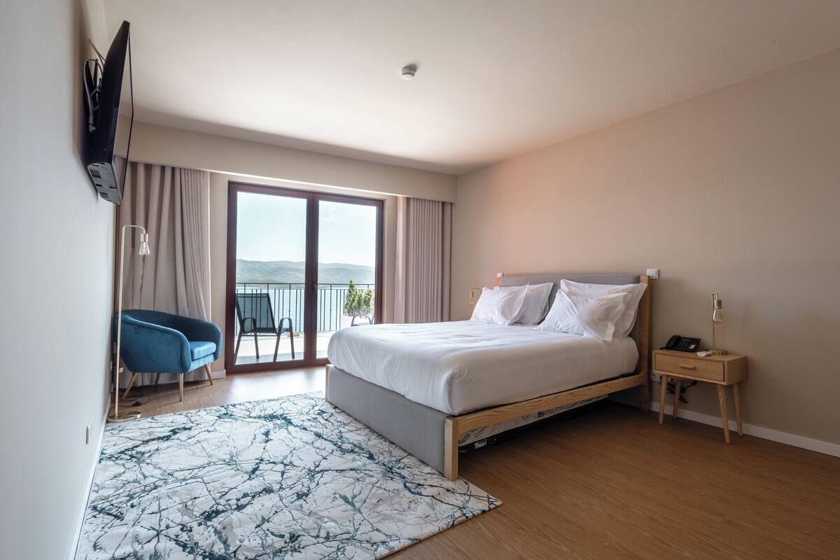 A modern hotel room with a large bed, a desk, a chair, and a view of the outdoors.