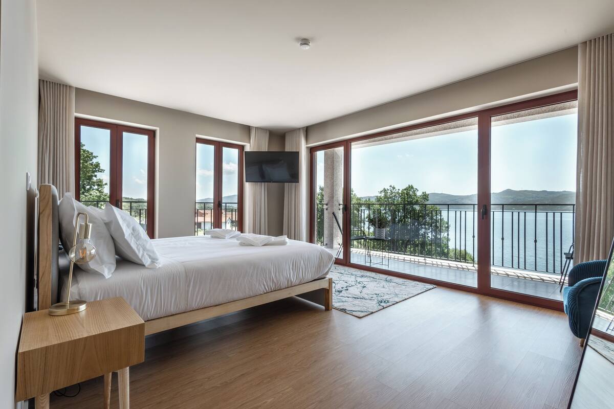 A modern bedroom with a large bed, a TV mounted on the wall, and a balcony with a view of the water.