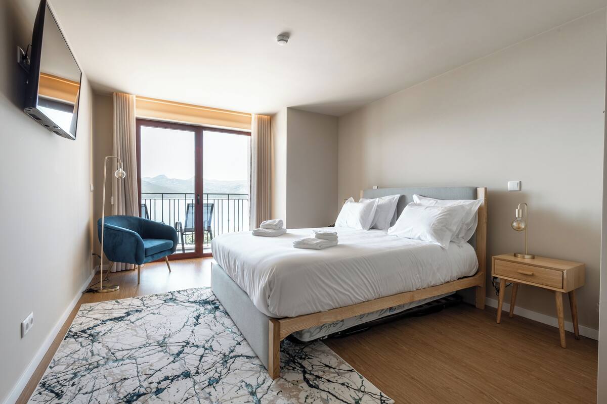 A modern hotel room with a large bed, a bedside table, a chair, and a view of the outdoors.