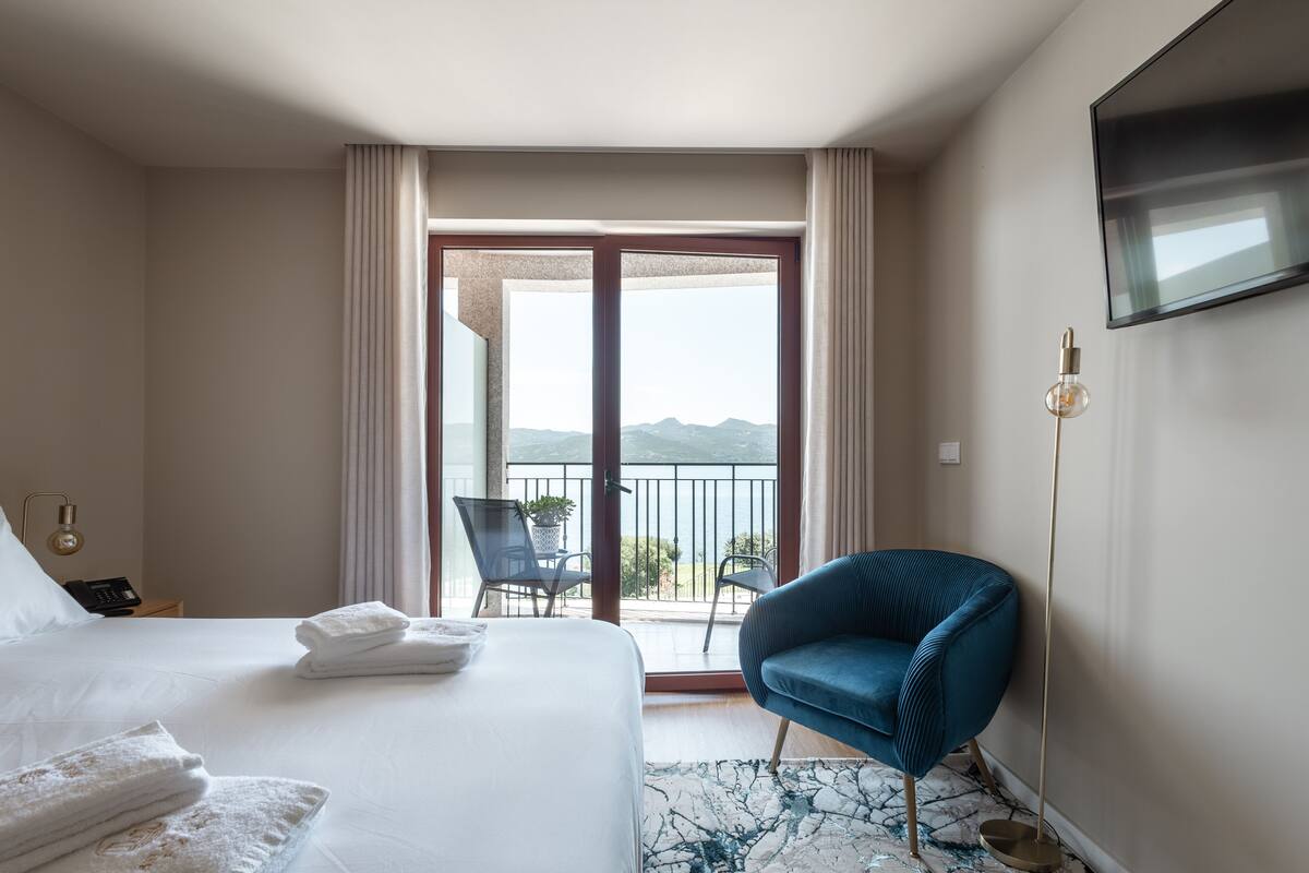 A hotel room with a bed, a blue chair, a TV, and a balcony with a view of the mountains.
