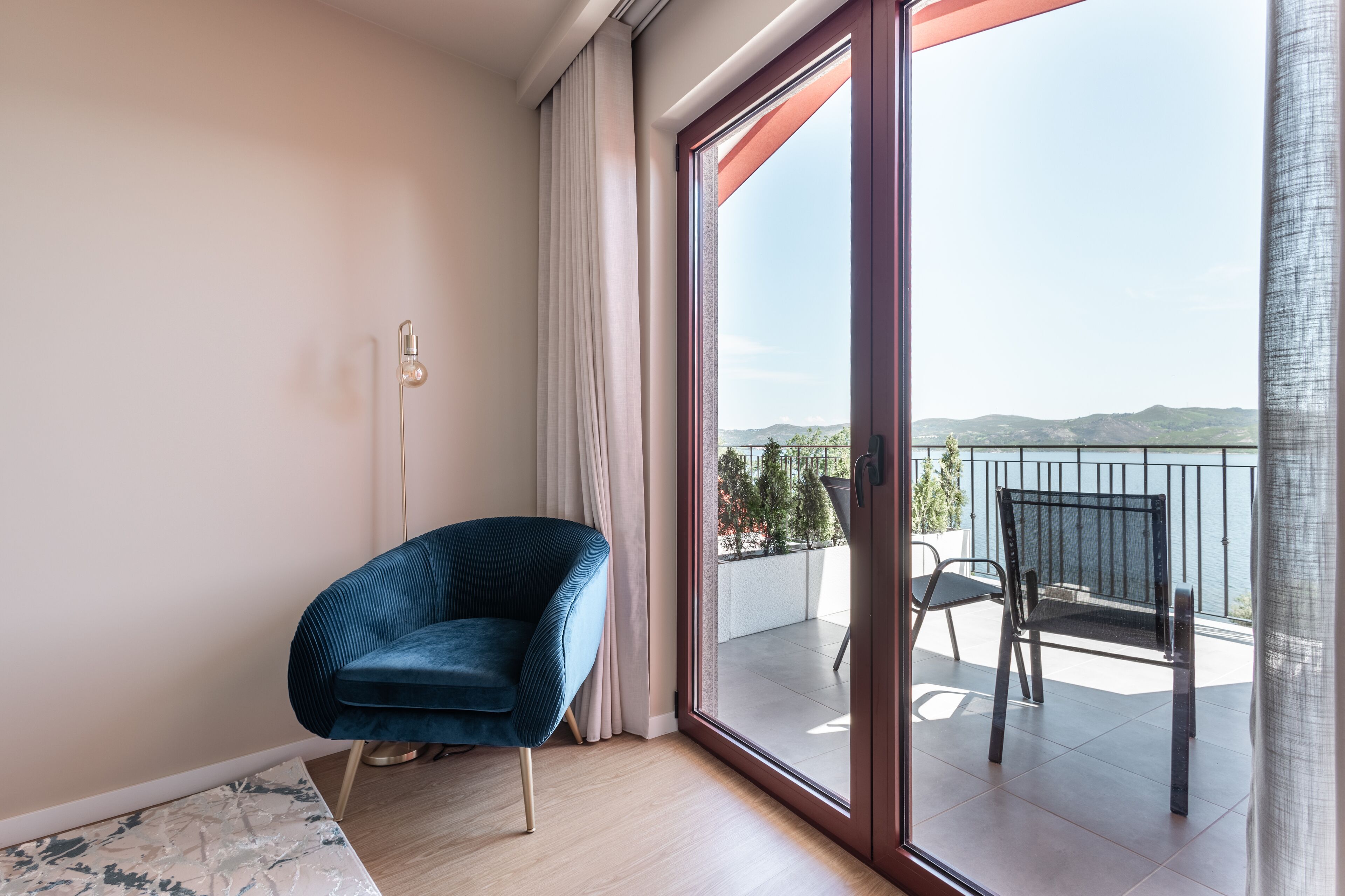 A modern room with a blue chair, a large sliding glass door leading to a balcony with a view of the sea and hills.