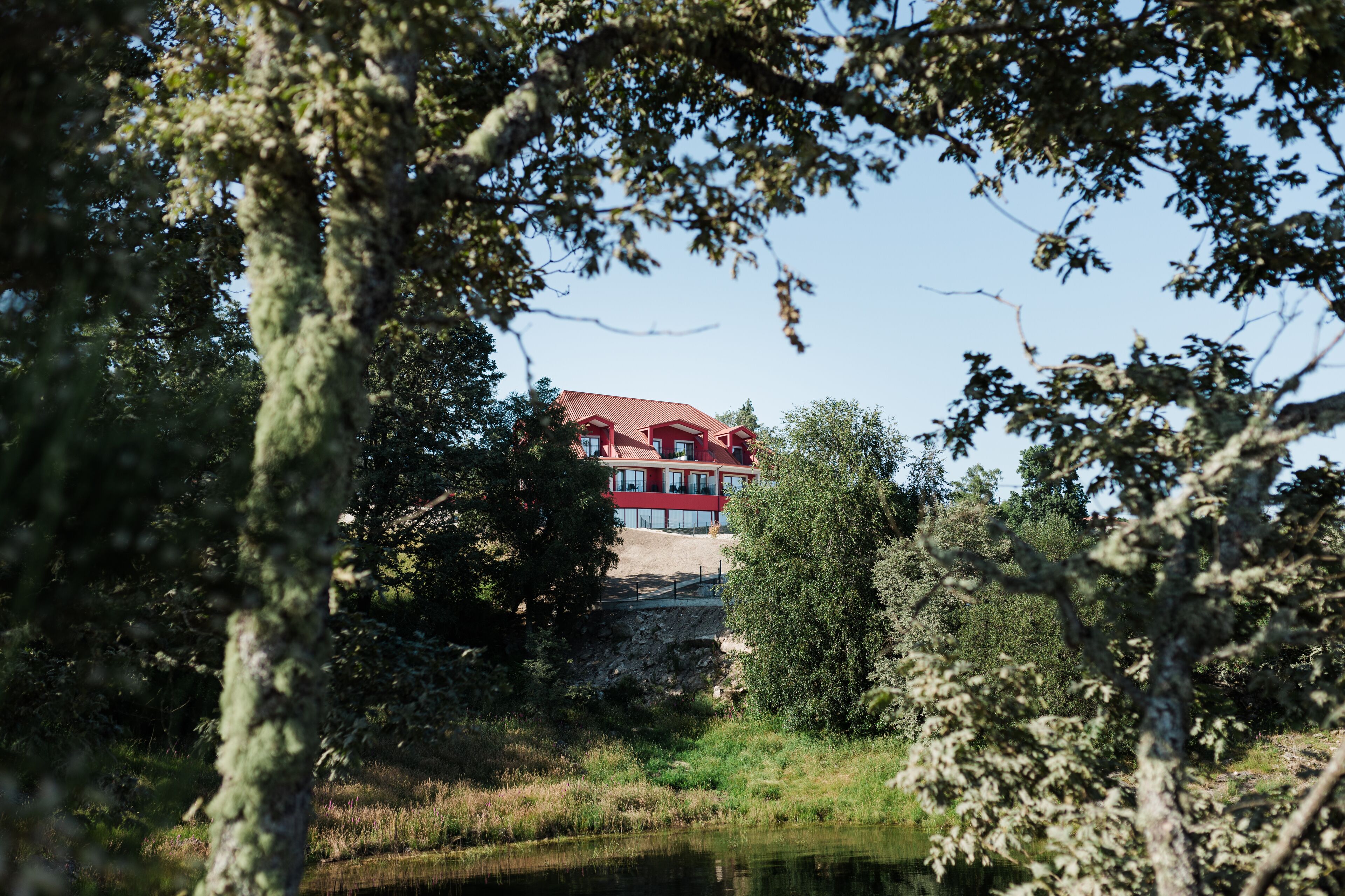 A red building with multiple windows and a sloped roof, surrounded by trees and a body of water.