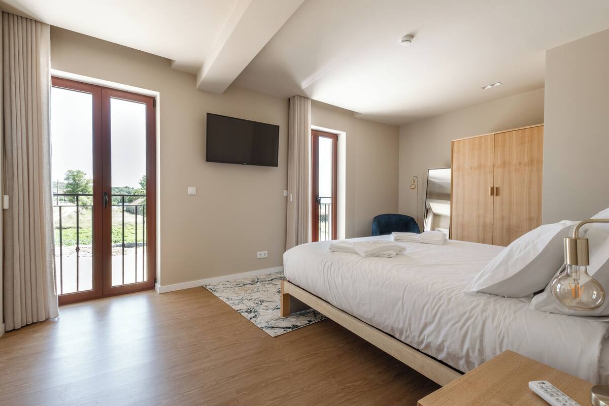 A modern hotel room with a large bed, a flat-screen TV, a wardrobe, and a balcony with a view.