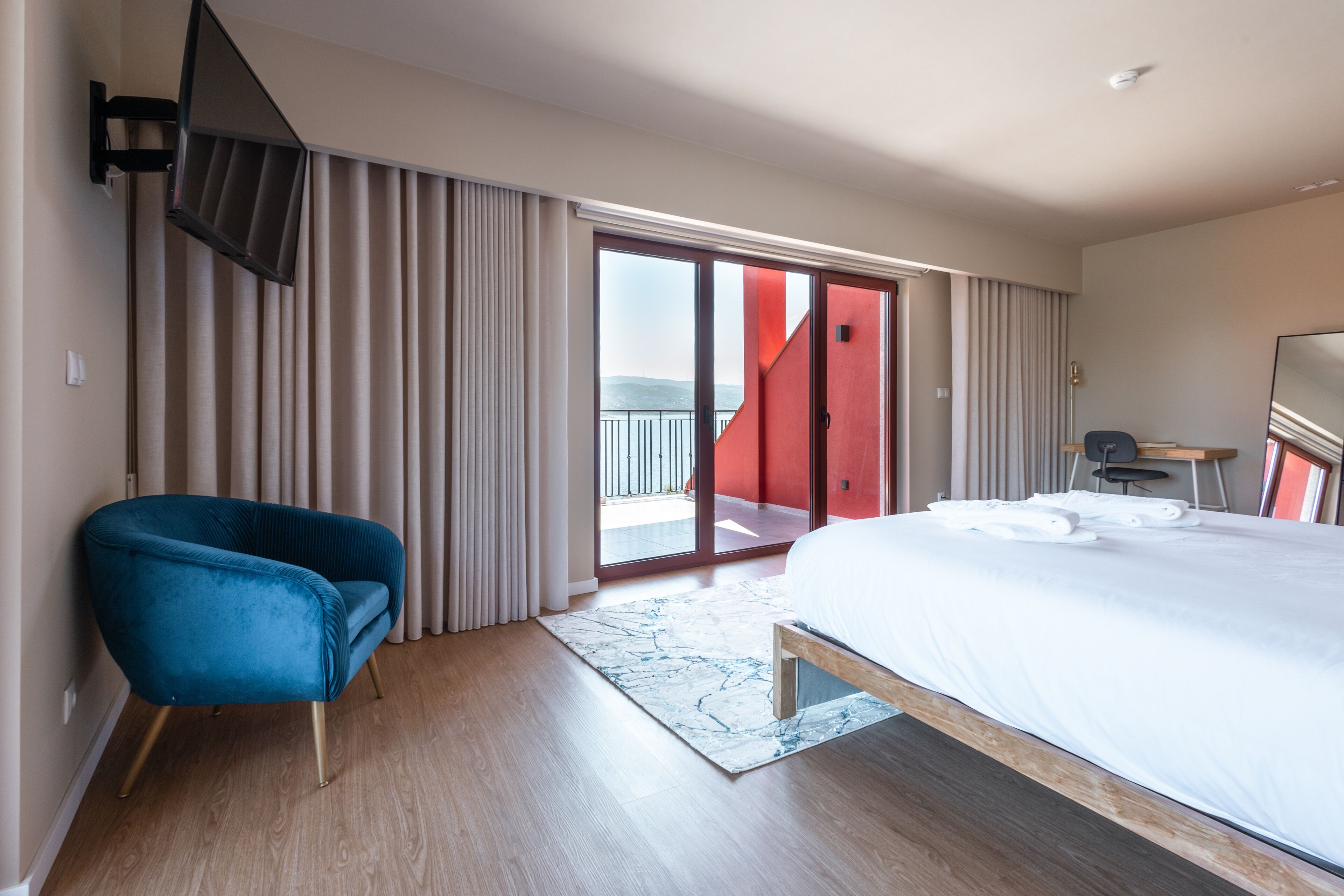 Hotellihuone, jossa on suuri sänky, sininen nojatuoli, puulattia ja näkymä lasiovesta.