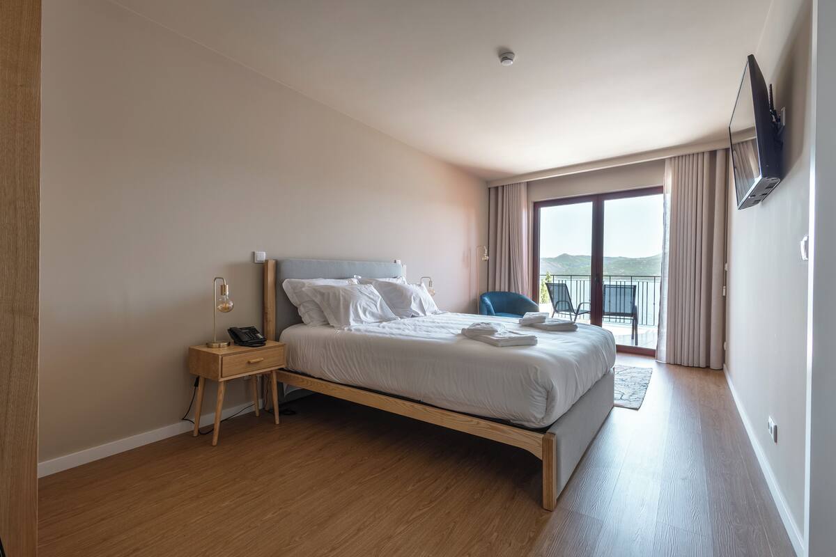 A modern hotel room with a wooden bed, a bedside table with a lamp, and a view of the outdoors through a large window.